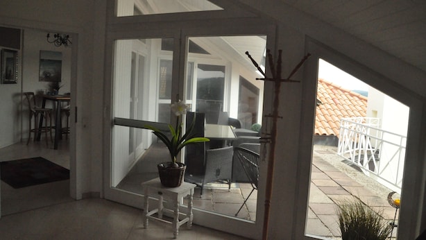 Quarto amplo com uma grande porta de vidro deslizante que dá para um espaço de pátio externo. No interior, há uma mesa branca com uma planta em vaso e um pequeno banquinho branco.