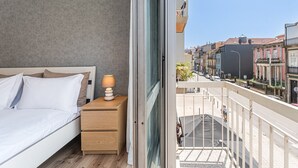A bedroom with a bed, bedside table, lamp, and a view of the city through a balcony. - GuestReady - Lovely family home in Boavista (Boavista)