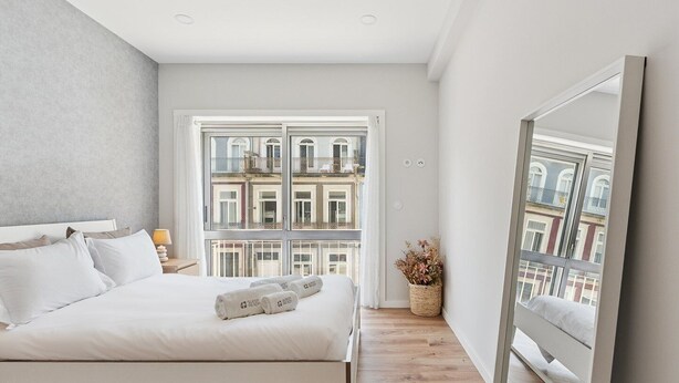A modern bedroom with a large bed, a nightstand, a lamp, and a view of the city through a large window. - GuestReady - Lovely family home in Boavista (Boavista)