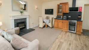 A living room with a fireplace, a sofa, a dining area, and a kitchen with wooden cabinets and appliances. - Midsummer Cottage (Malton)