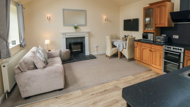 A spacious living room with a fireplace, a dining area, and a kitchen with wooden cabinets and modern appliances. - Midsummer Cottage (Malton)