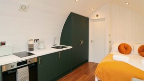Una habitación de hotel compacta con kitchenette, una cama con almohadas naranjas y piso de madera.