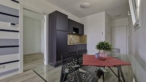 A modern dining area with a glass table and black chairs, a small kitchen with dark cabinets, and a potted plant on the table.