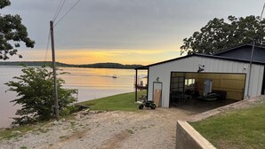 A gravel driveway leading to a building with a covered outdoor area, a boat on the water, and a sunset in the background. - Lake Front House with Private Dock and boat rental available!  Lake Eufala (Checotah)