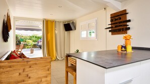 A modern kitchen with a large island, wooden bench seating, and a view of the outdoors through a sliding door. - Pymen - Cocon climatisé avec terrasse et jardin (Rivière-Salée)