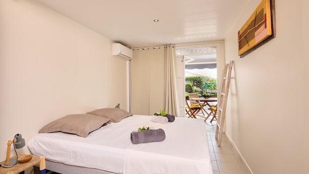 A bedroom with a bed, a small table with a lamp, and a view of an outdoor area with a table and chairs. - Pymen - Cocon climatisé avec terrasse et jardin (Rivière-Salée)