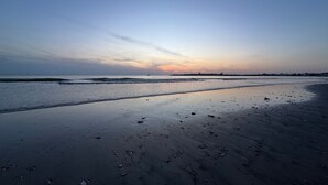 Una playa con arena mojada y olas suaves, cielo despejado y edificios a lo lejos.