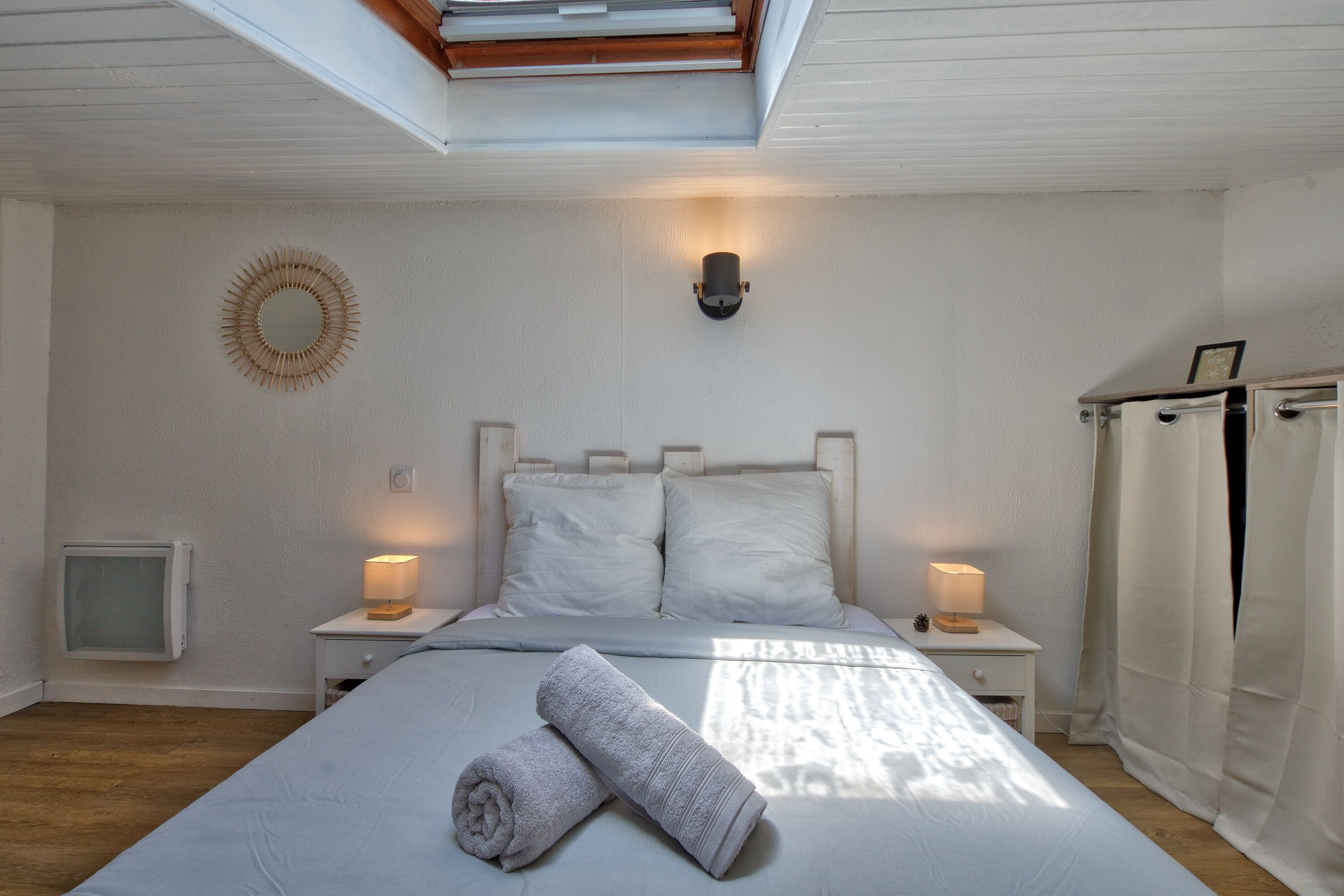 Ein Schlafzimmer mit Dachfenster, zwei Nachttischlampen, einem Bett mit weißen Leinen, einem zusammengerollten Handtuch, einem kleinen Nachttisch und einer Wandleuchte.
