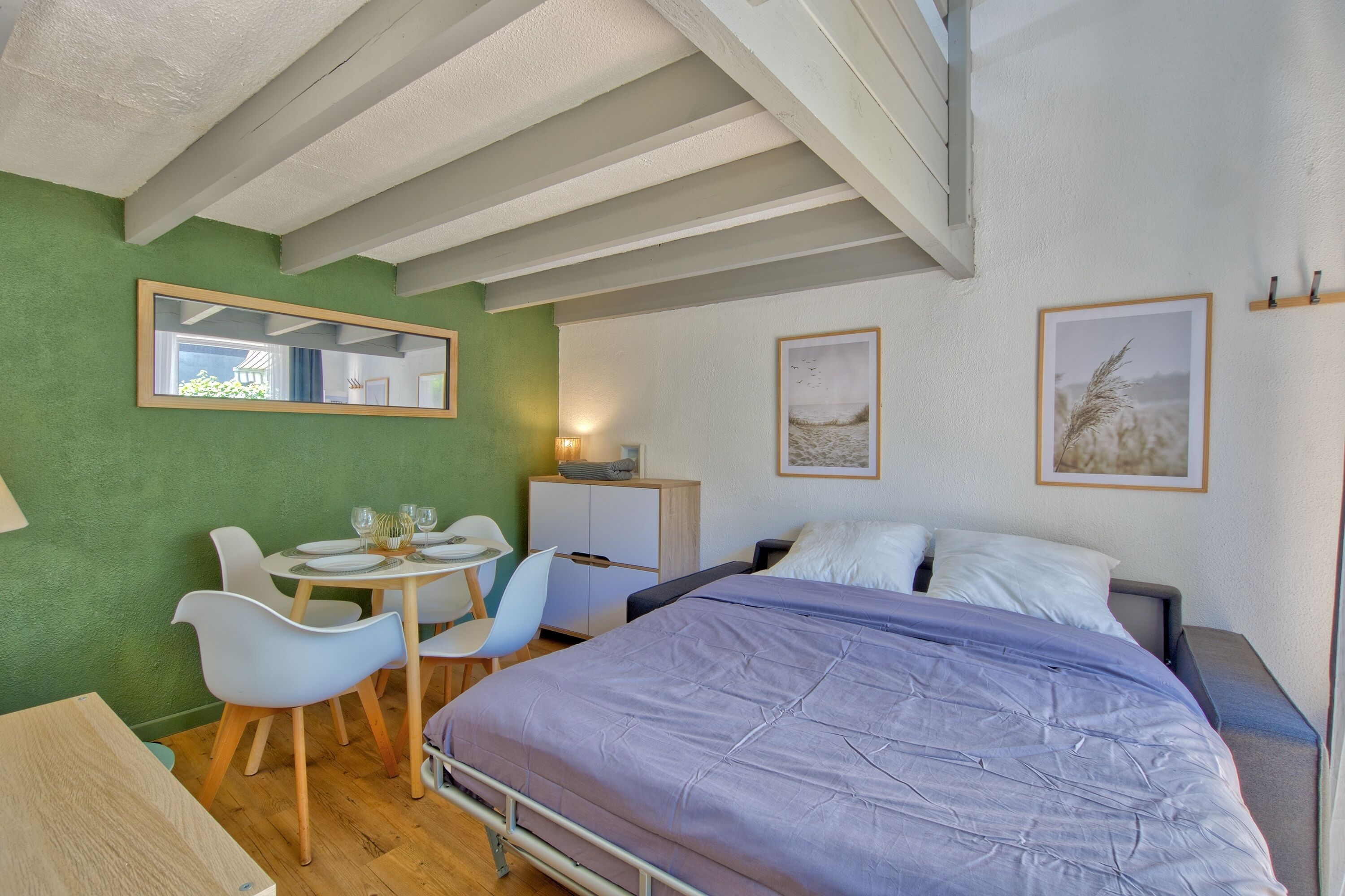 A bedroom with a bed, a desk with chairs, a mirror, and two framed pictures on the wall.