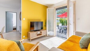 A modern living room with a yellow accent wall, a flat-screen TV on a wooden console, a sofa with yellow cushions, and a sliding glass door leading to an outdoor area with a red umbrella.