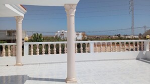Une terrasse sur le toit, agrémentée de balustrades et de colonnes blanches, un ciel dégagé et des bâtiments qui se dressent à l’horizon.