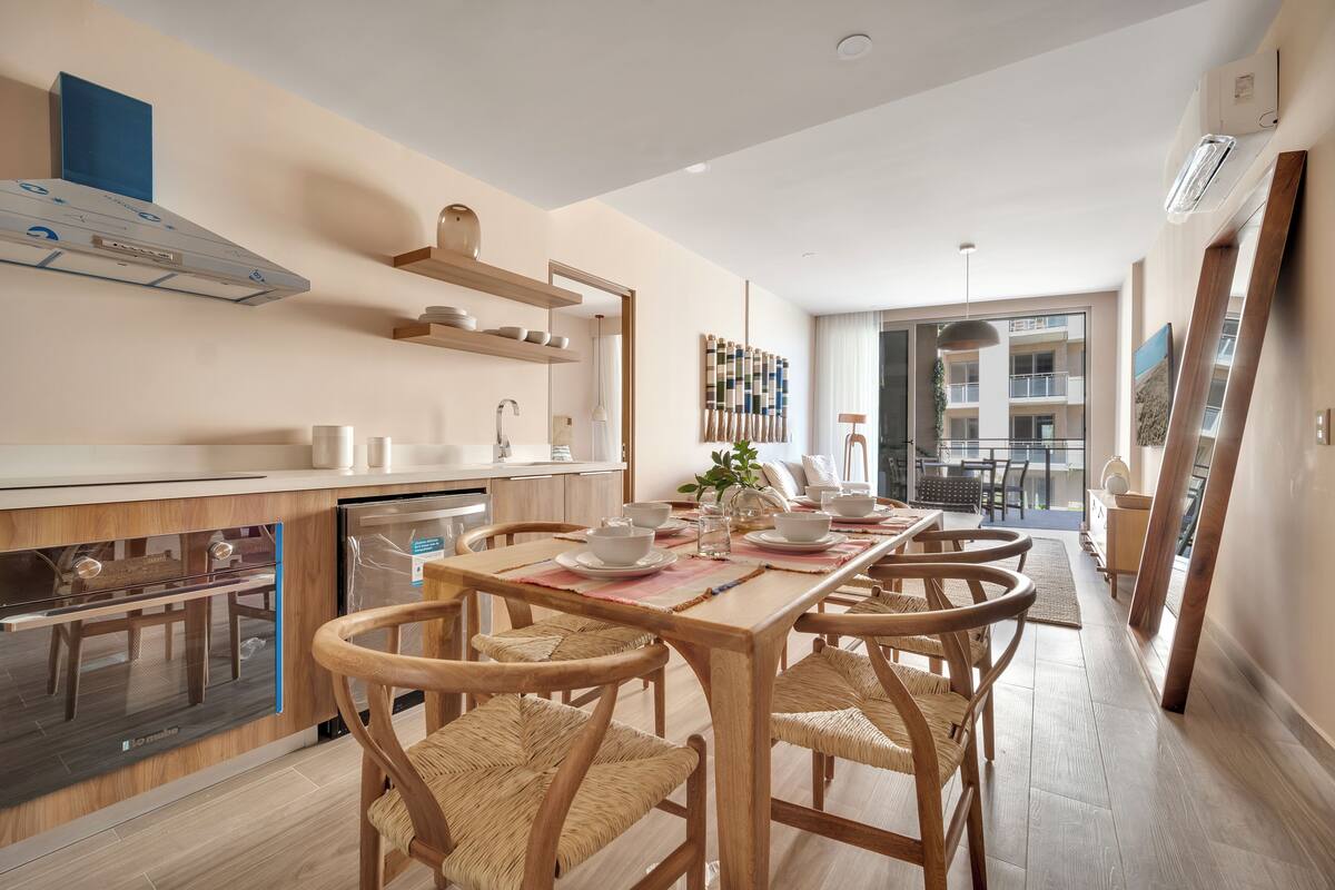 A modern kitchen with built-in appliances, a dining area with wooden chairs and a table set for four, and a view of the living area through an open door.