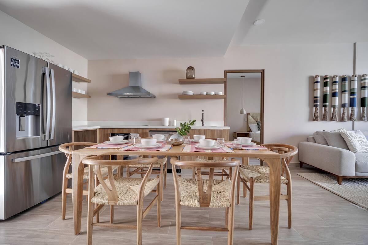 A modern kitchen with a built-in refrigerator, wooden dining table with chairs, and a sofa in the living area.