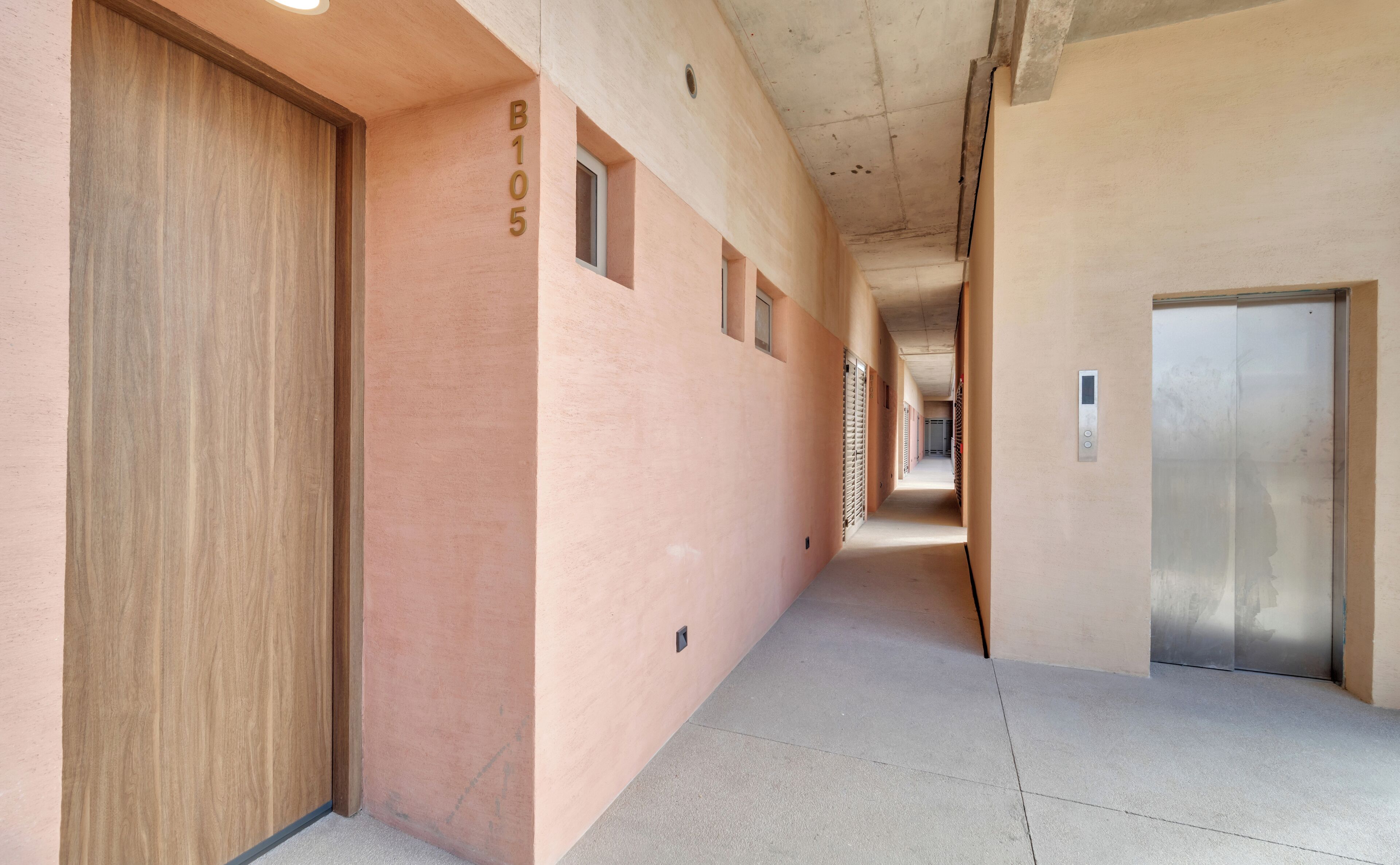 Un pasillo con una puerta de madera, una pared con el número B105 y una puerta de ascensor metálica.