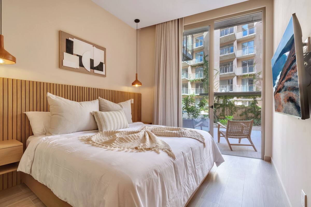 A bedroom with a wooden headboard, a large bed with white bedding, a bench, a hanging pendant light, a framed abstract artwork, and a balcony with a view of a building and greenery.
