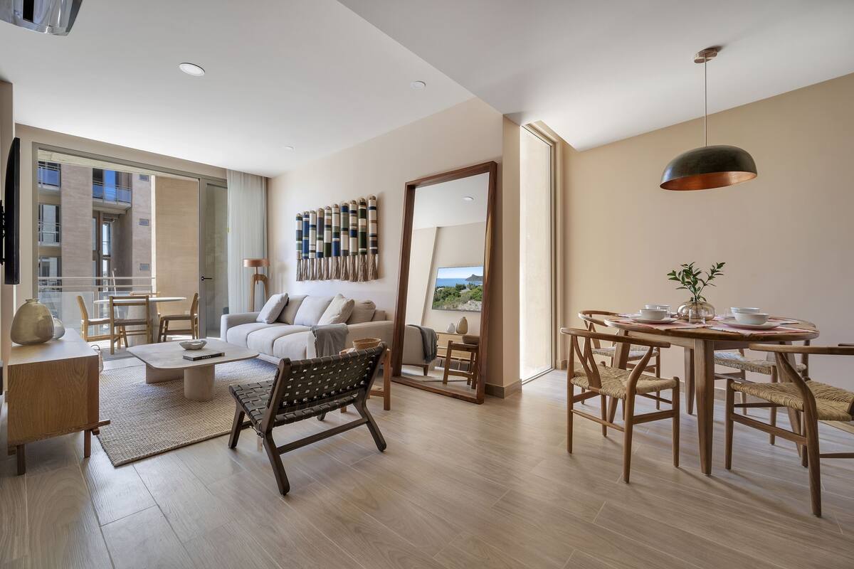 A modern living room with a sofa, coffee table, dining table, and chairs. There is a large mirror and a hanging light fixture.