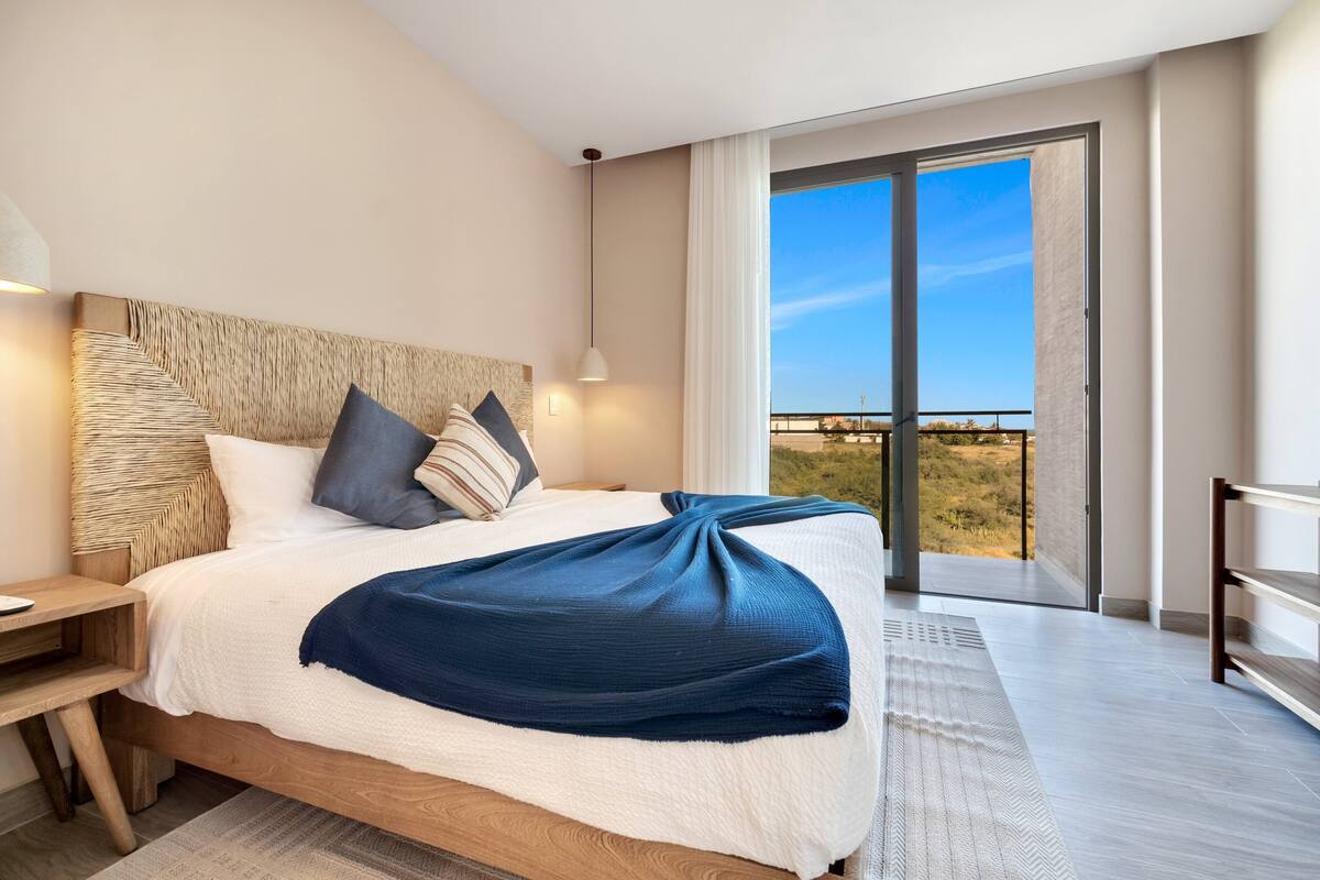 A modern hotel room with a large bed, a nightstand, a desk, and a balcony with a view.
