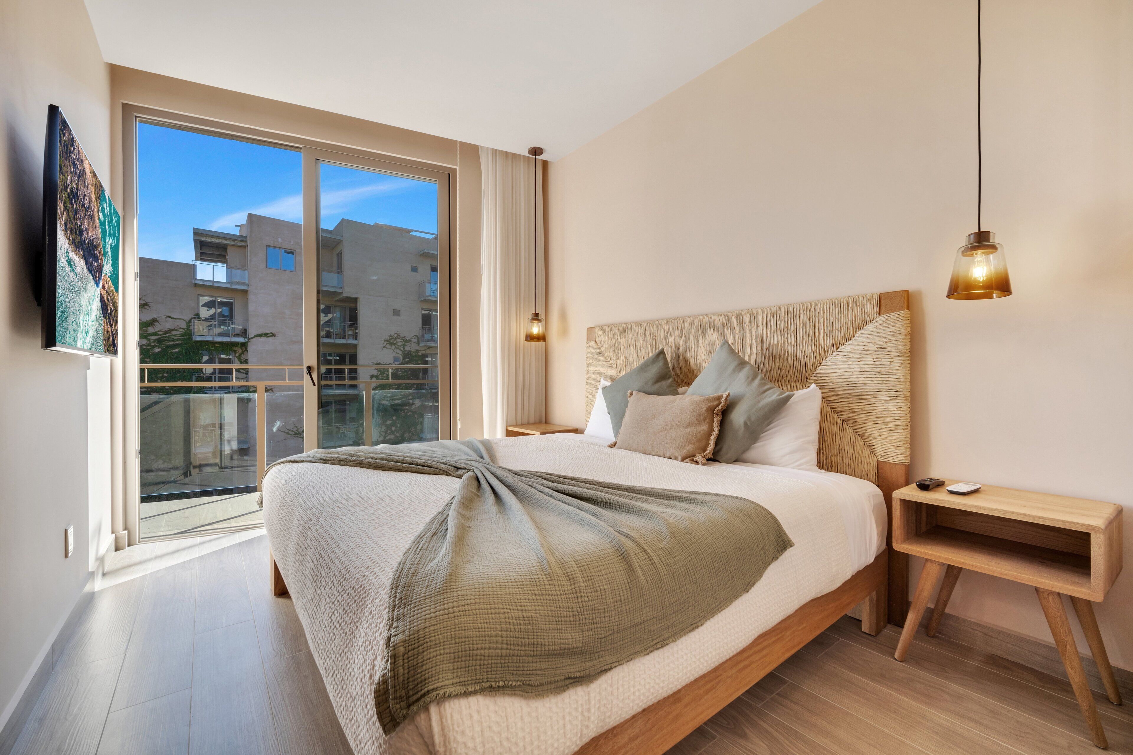Une chambre moderne avec un grand lit, une table de chevet en bois, une lampe suspendue et une vue sur les immeubles par une grande fenêtre.