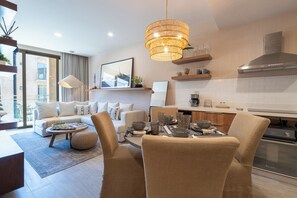 A modern living room with a sofa, chairs, a dining table set, a large window, and a kitchen area with a stove and cabinets.