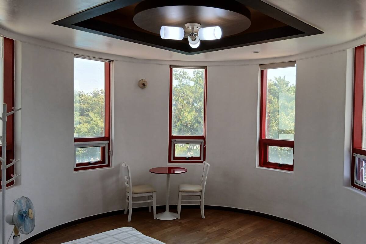 Ein Eckzimmer mit Holzboden, Deckenventilator, kleinem Tisch mit zwei Stühlen und großen Fenstern mit roten Rahmen.