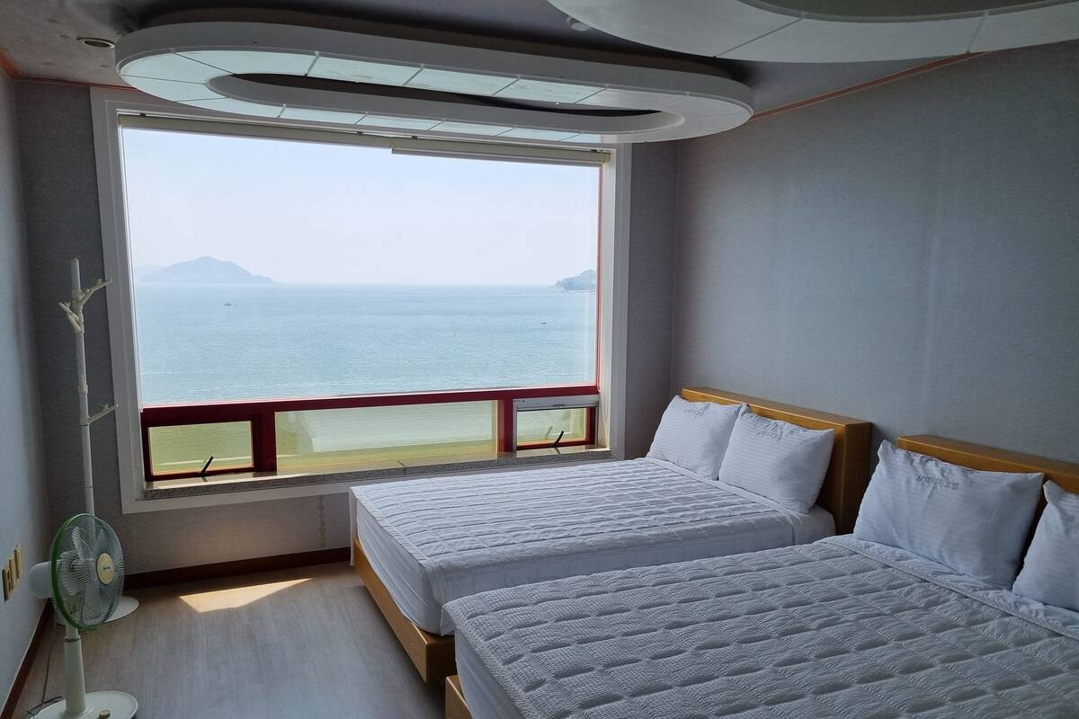 Ein Hotelzimmer mit zwei Betten, einem großen Fenster mit Meerblick, einem Standventilator und einem Holzboden.