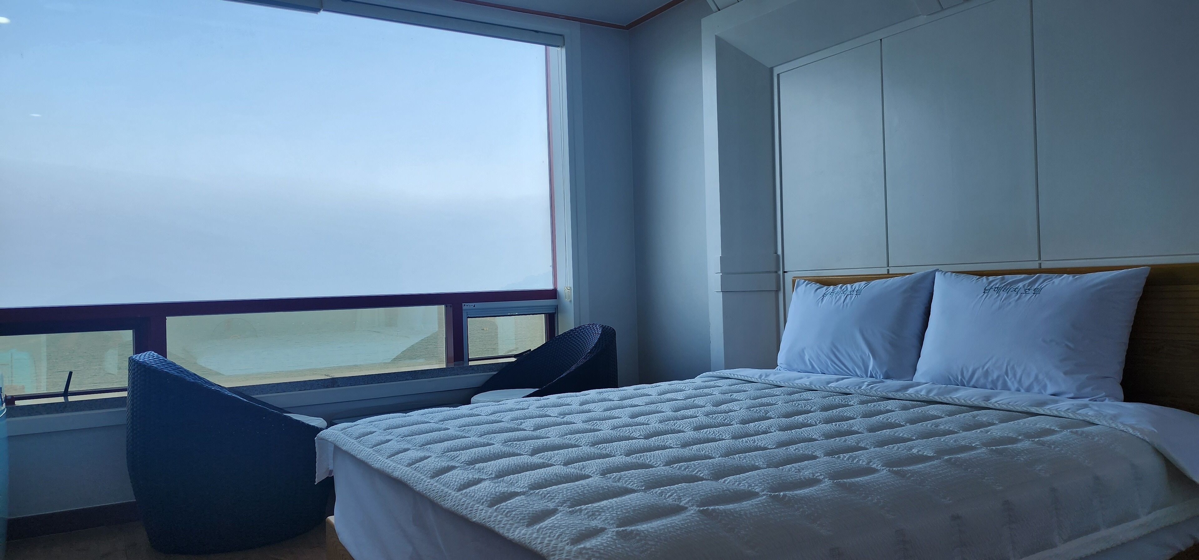 Une chambre d’hôtel avec une grande fenêtre, un lit avec une literie blanche, une tête de lit en bois et une chaise de couleur foncée.