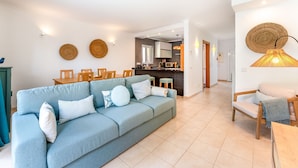 A modern living room with a light blue sofa, wooden chairs, and a dining table. There is a kitchen area with a bar and a hanging woven light fixture.