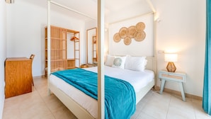 A four-poster bed with a teal bedspread, a wooden wardrobe, a bedside table with a lamp, and a wall with woven art.