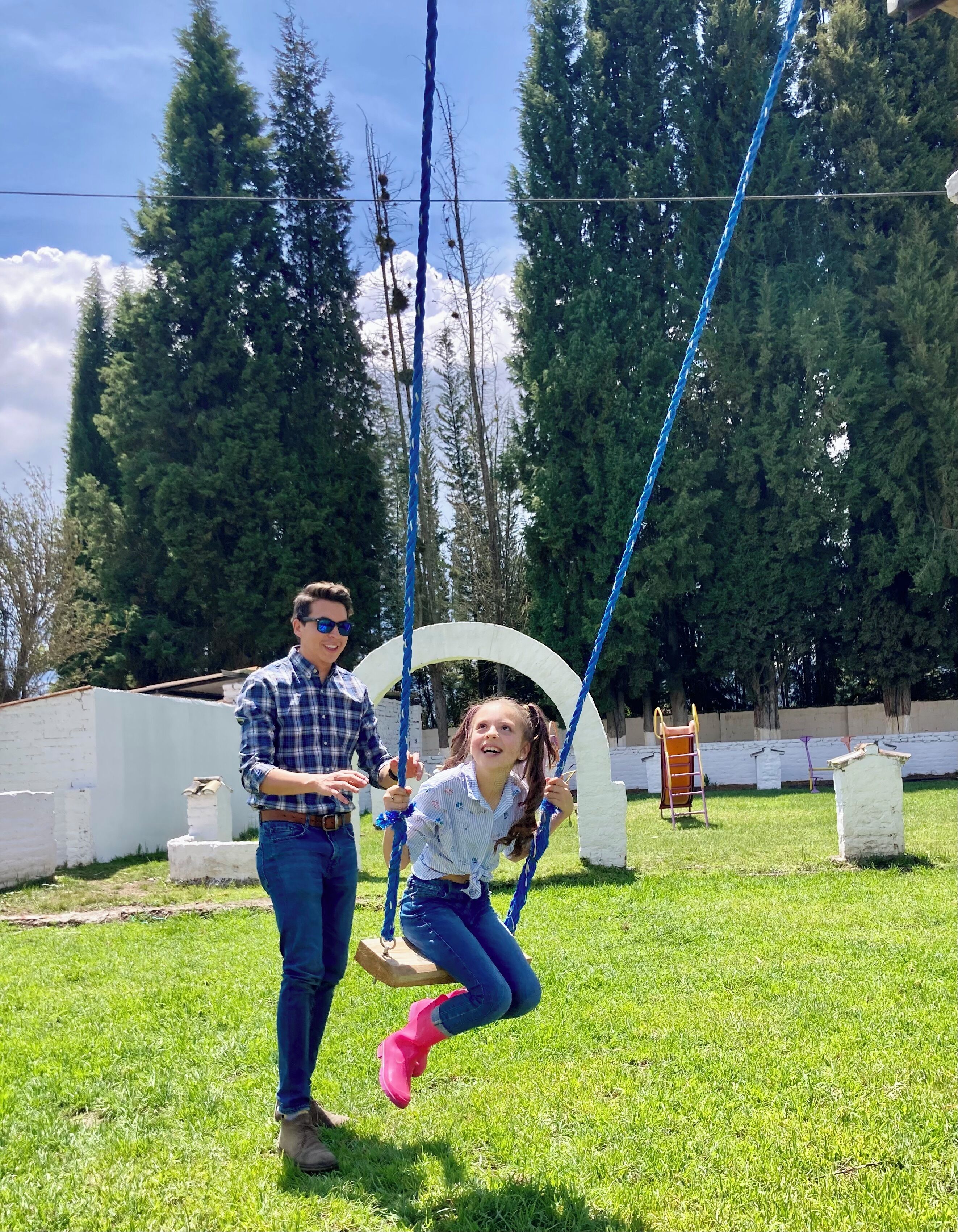 A grassy outdoor area with a swing set, a white wall, and trees in the background.