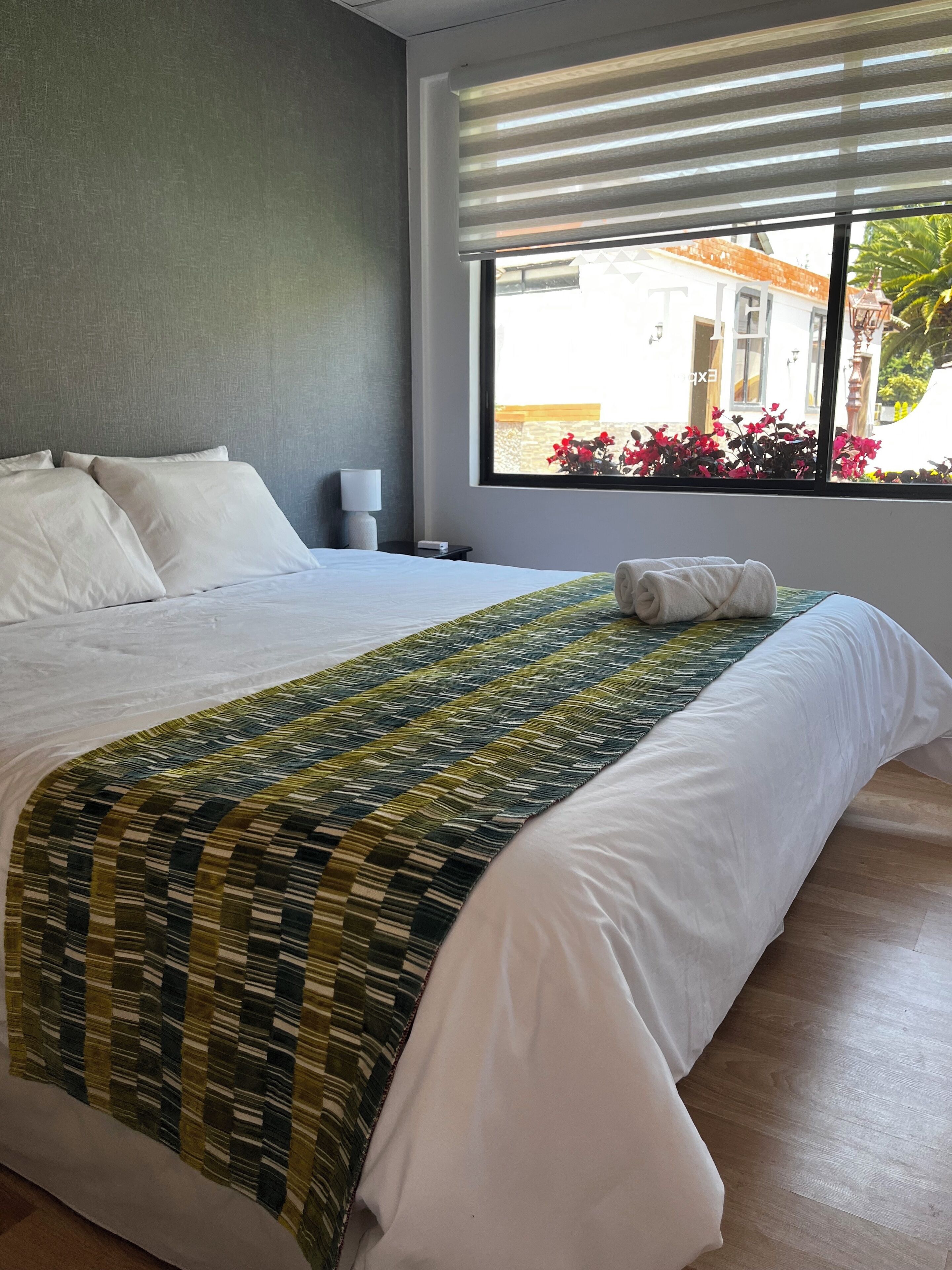 Habitación de hotel con una cama grande, una ventana con persianas y vistas a edificios y flores.
