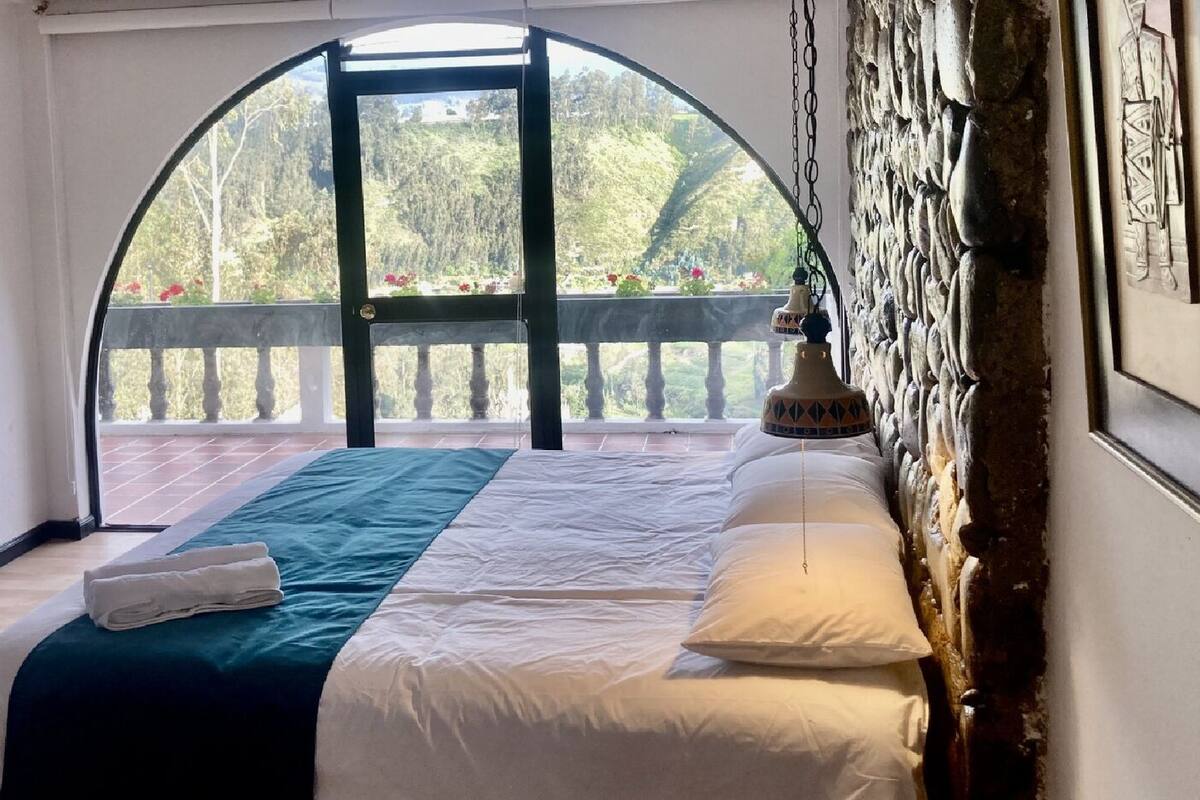 Ein Schlafzimmer mit einem großen Bogenfenster, einem Bett mit weißer Bettwäsche, einer türkisblauen Tagesdecke und einem kleinen Tisch mit einem dekorativen Gegenstand.