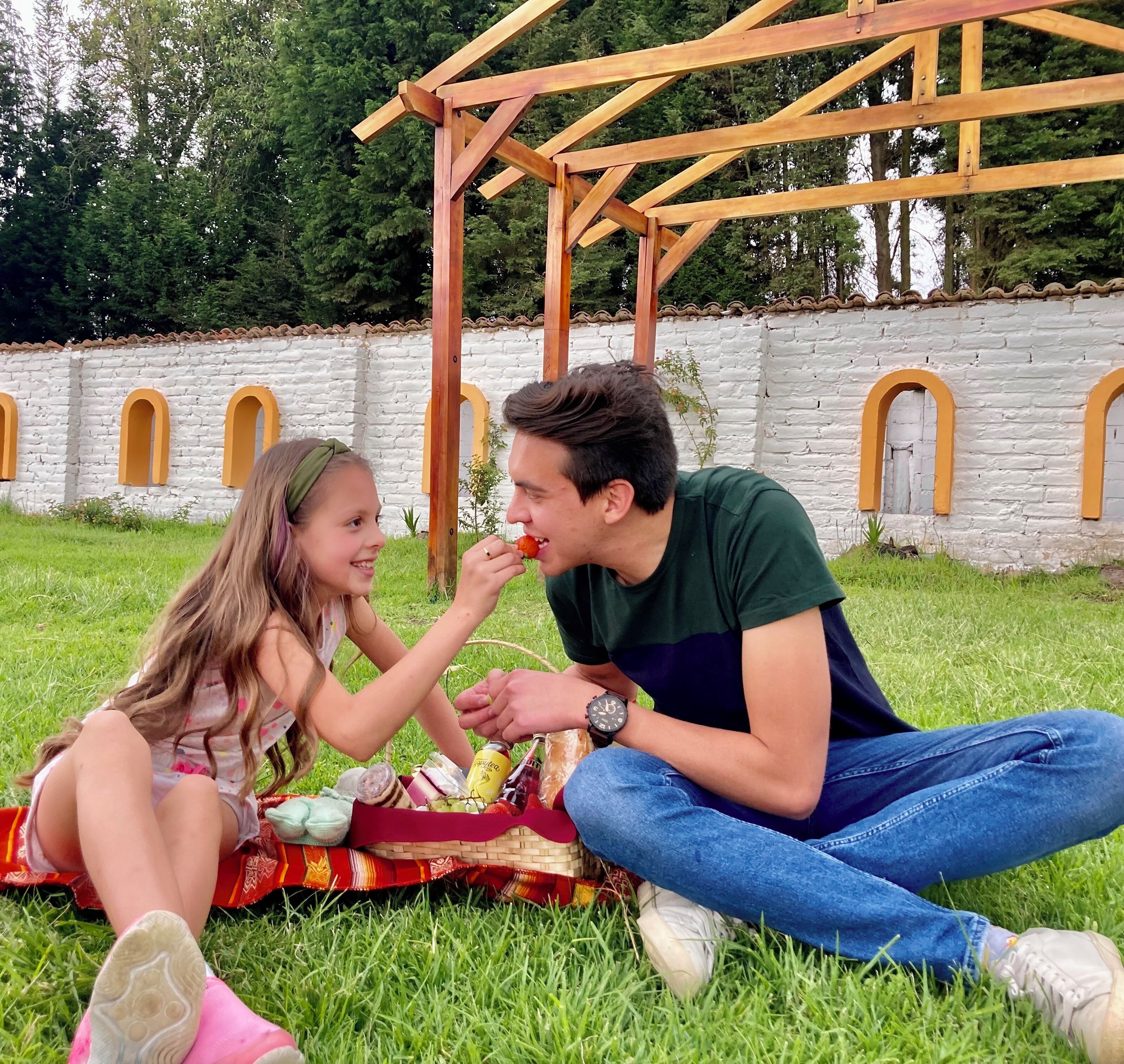 Una pareja sentada sobre la hierba con una preparación para picnic, incluyendo una cesta con comida y bebidas, frente a un muro de piedra con ventanas arqueadas.