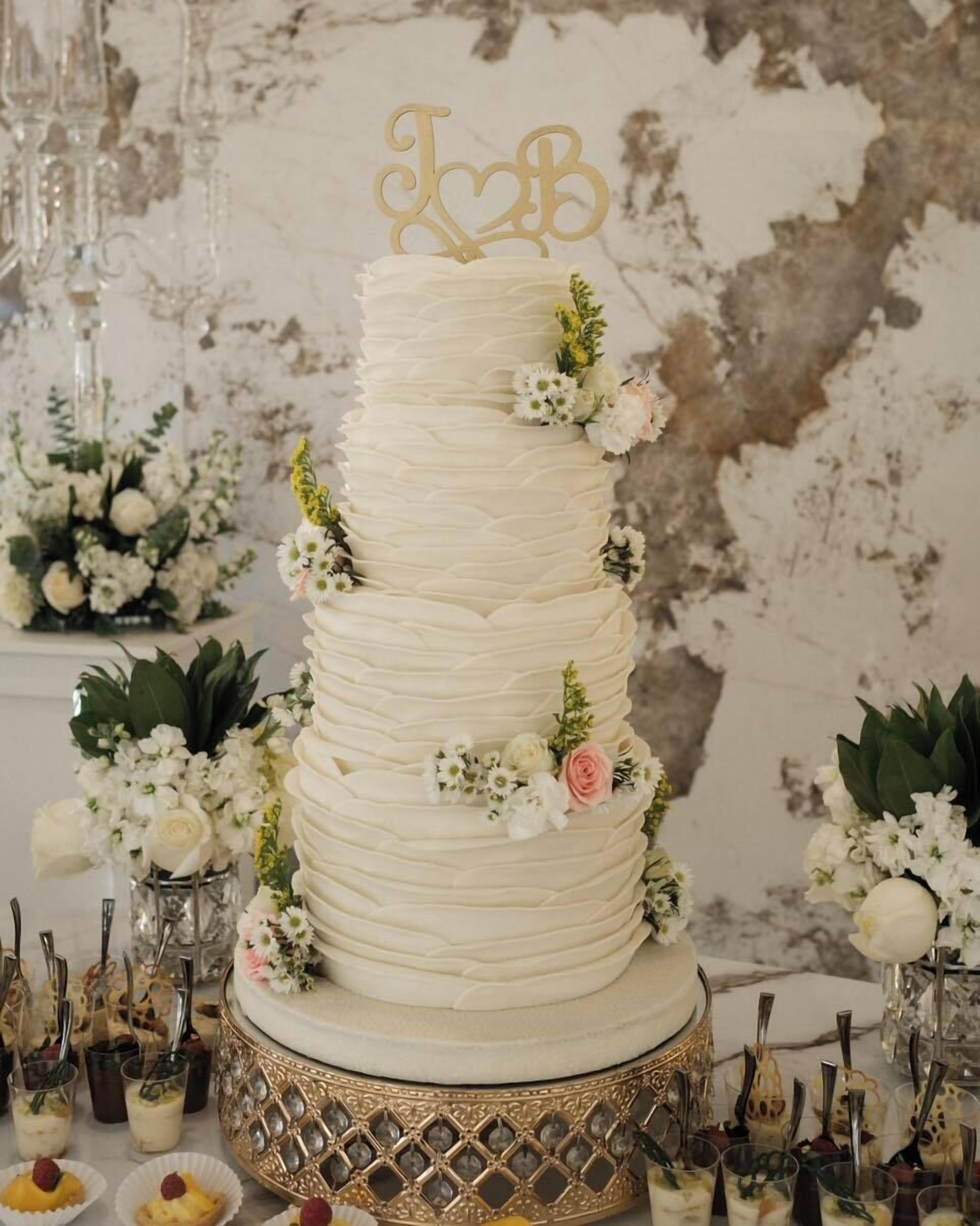 A multi-tiered wedding cake with a 'T&B' topper, surrounded by floral arrangements and dessert cups.