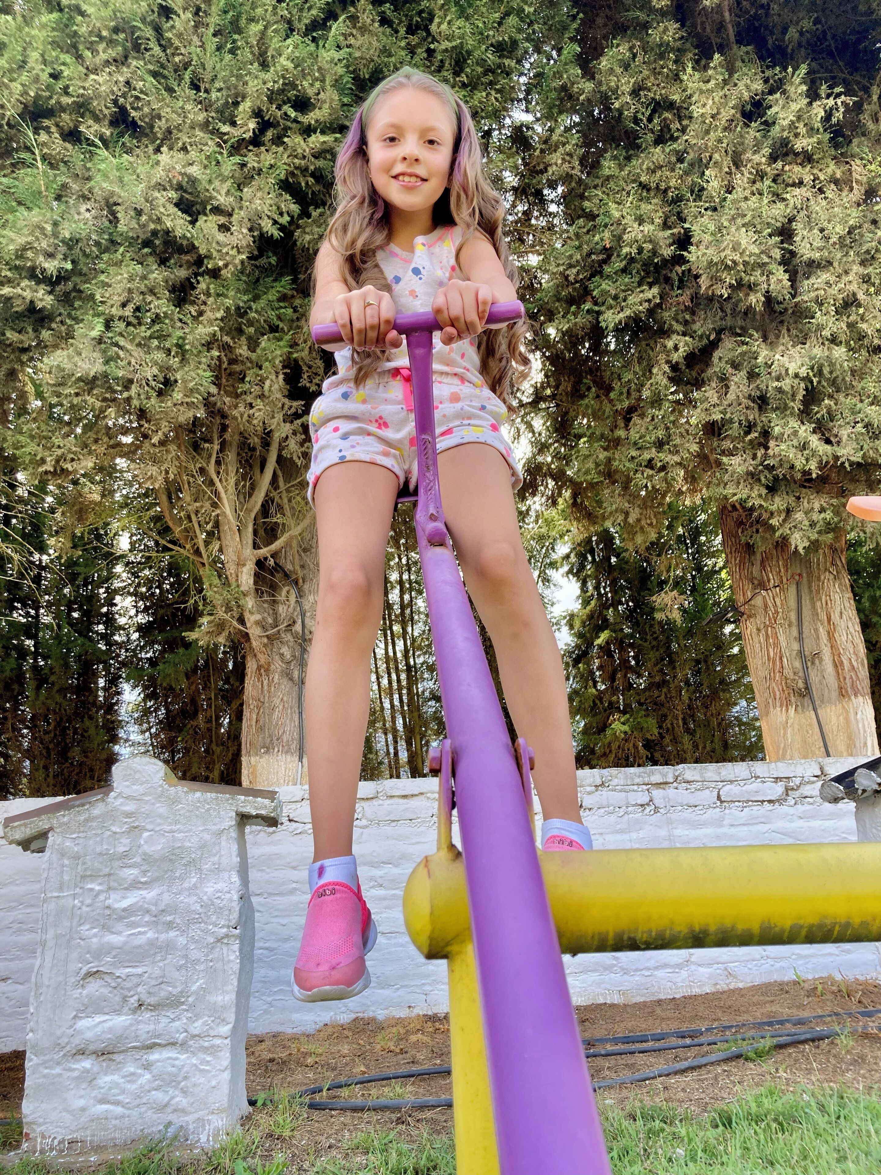 Un nen o nena en un seesaw de morat i groc a un parc, amb arbres i una paret de pedra al fons.