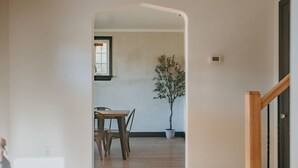 A hallway leading to a dining area with a wooden table and chairs, a potted plant, and a window with blinds. - The Safe Harbor Home | Quiet Retreat, Fire Pit, Nature (Conestoga)