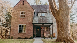 A two-story brick house with a gabled roof, a front porch, and a large tree in the foreground. - The Safe Harbor Home | Quiet Retreat, Fire Pit, Nature (Conestoga)