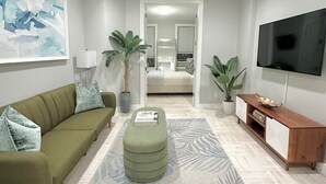 A modern living room with a green sofa, a round ottoman, a wooden TV stand, and a large abstract painting on the wall.