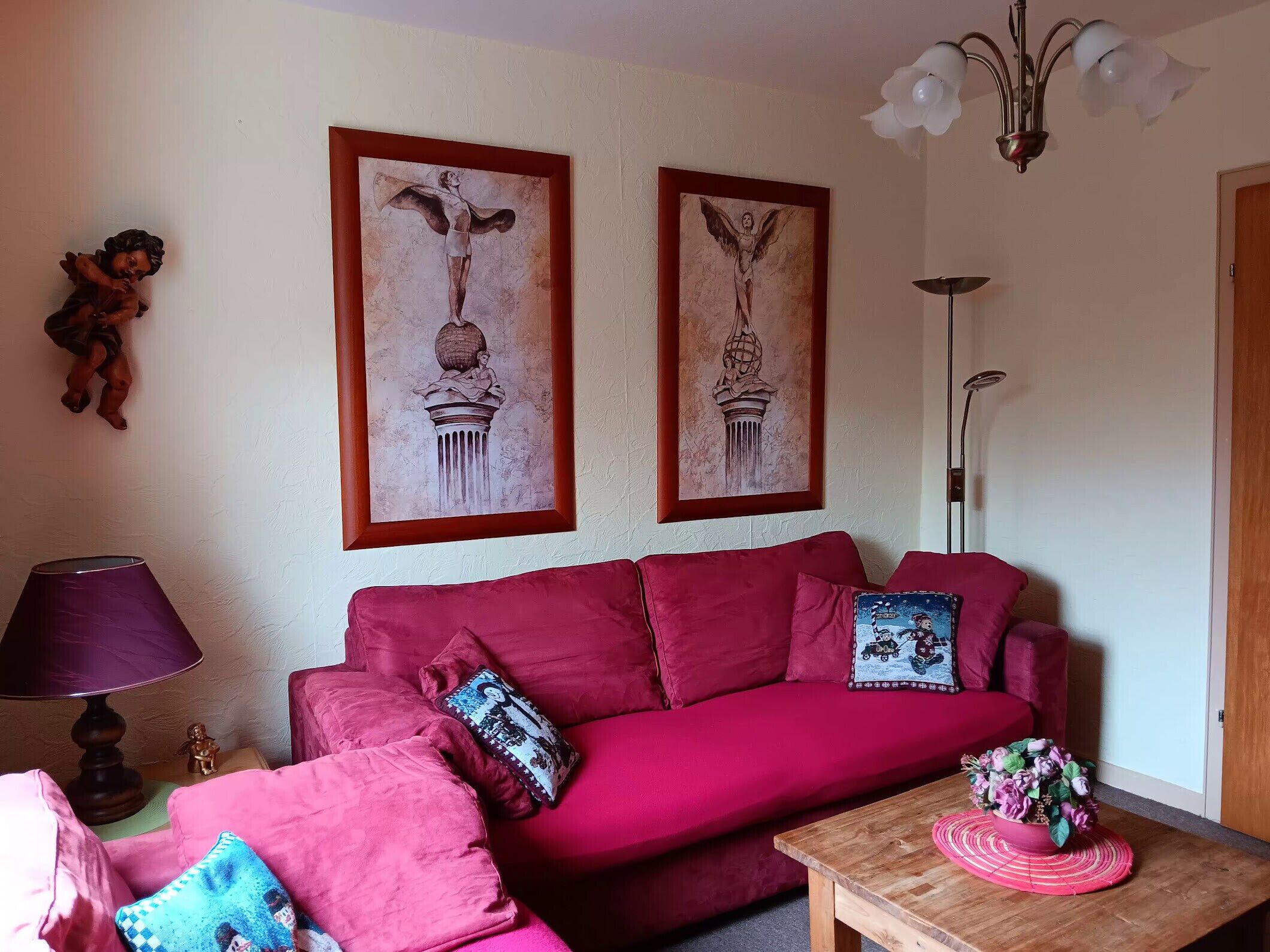 Een woonkamer met een rode bank, een bijzettafeltje met een bloemstuk, en twee kunstwerken in lijsten aan de muur.
