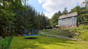A grassy backyard with a trampoline, a green slide, and a house in the background.