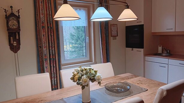 A dining area with a table, chairs, a vase with flowers, a clock, and a window with curtains.