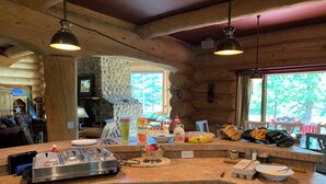 A kitchen with a stone fireplace, wooden beams, and a large island with various food items and kitchen appliances. - The Lake House (Rimbey)