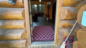 A log cabin interior with a wooden floor, a red patterned mat at the entrance, and a broom leaning against the wall. - The Lake House (Rimbey)