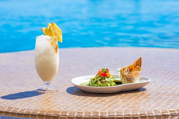 Un verre contenant une boisson crémeuse, agrémentée d’une garniture, une assiette avec une salade et un élément plié, et une piscine en arrière-plan.