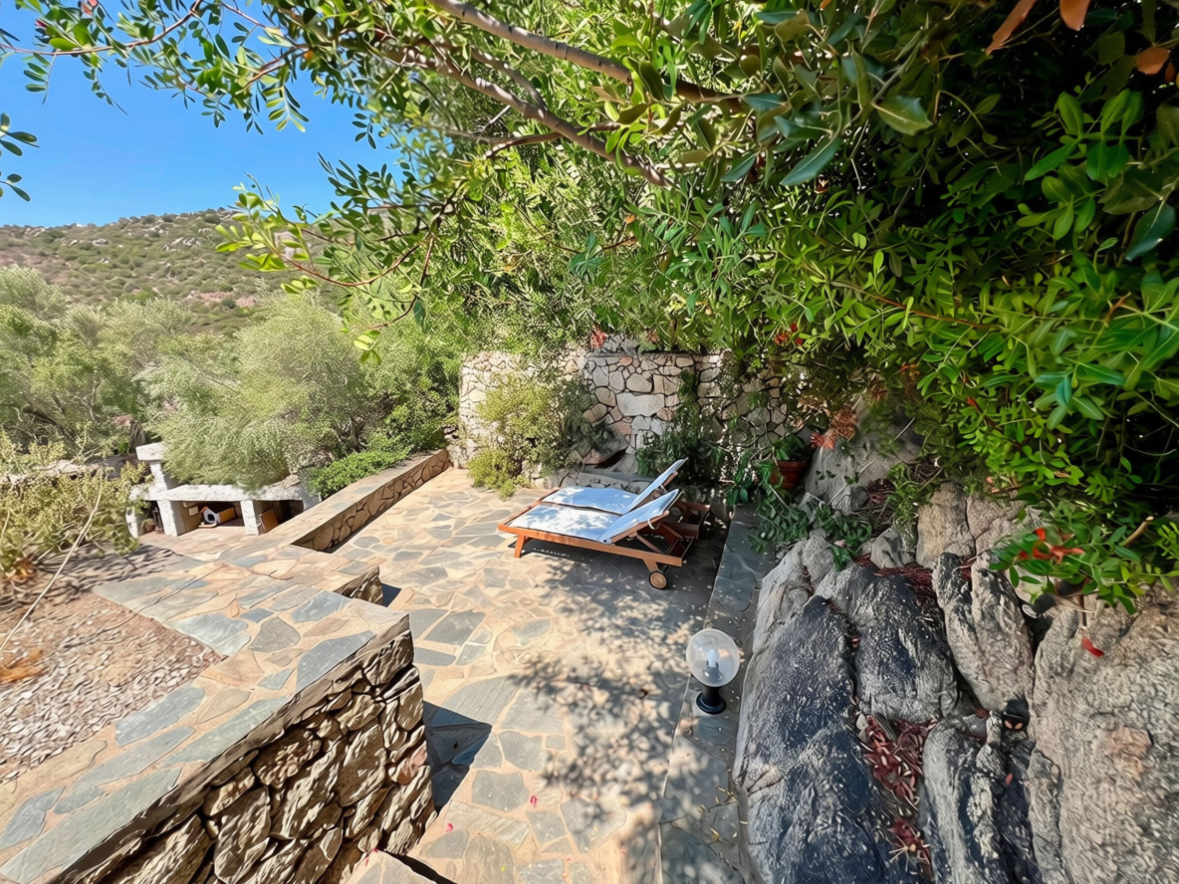 A stone patio with a lounge chair and a table, surrounded by greenery and a stone wall.