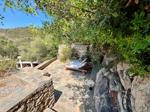 A stone patio with a lounge chair and a table, surrounded by greenery and a stone wall.