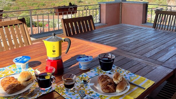 A wooden table set with breakfast items, including croissants, coffee, and a thermos, on a balcony with a view of a green hillside. - Villa Olbia surrounded by greenery.Perfect mix of peace and the sea Sardegna (Osseddu)
