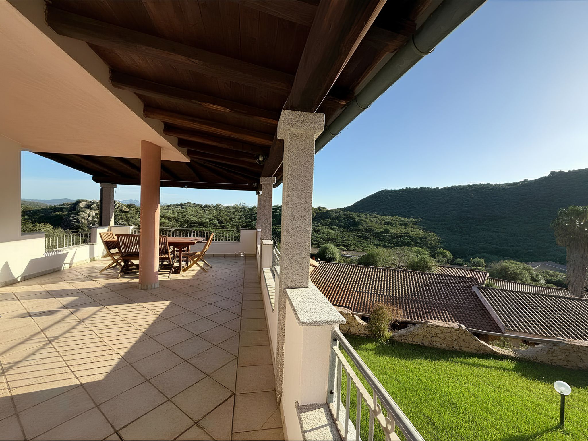 A covered outdoor area with a tiled floor, wooden furniture, and a view of a mountainous landscape.