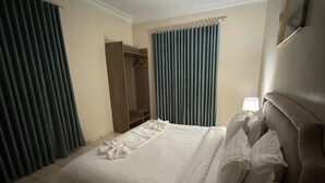 Habitación de hotel con una cama grande, un armario, una mesita de noche con lámpara y una ventana con cortinas.