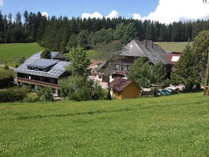 Une maison de campagne avec un toit en pente, entourĂ©e dâun champ verdoyant et dâune colline boisĂ©e.