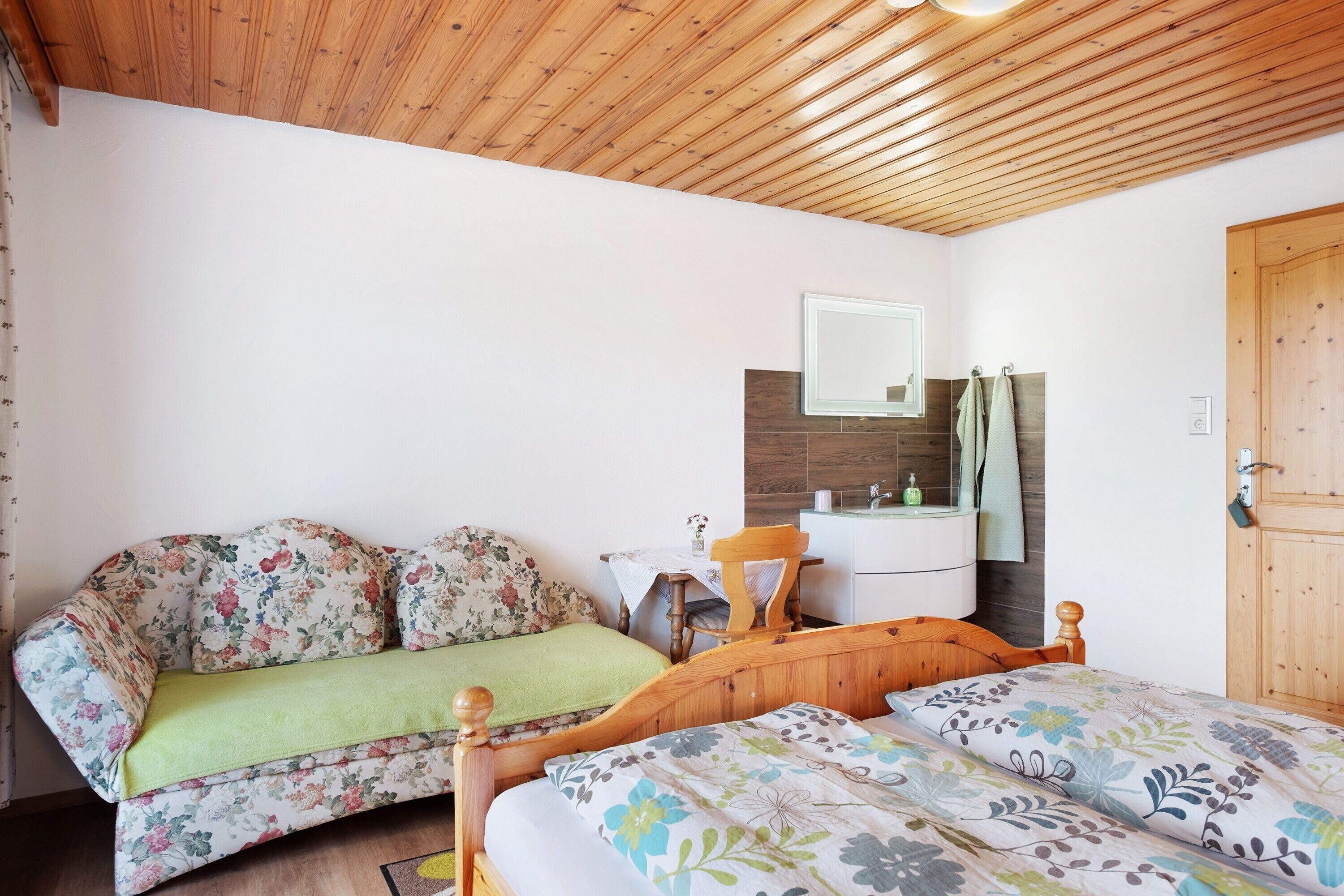 Ein Schlafzimmer mit Holzboden, einem Bett mit floralem Bettzeug, einem Holzstuhl, einem kleinen Tisch mit einer Vase und einem Holzschrank.